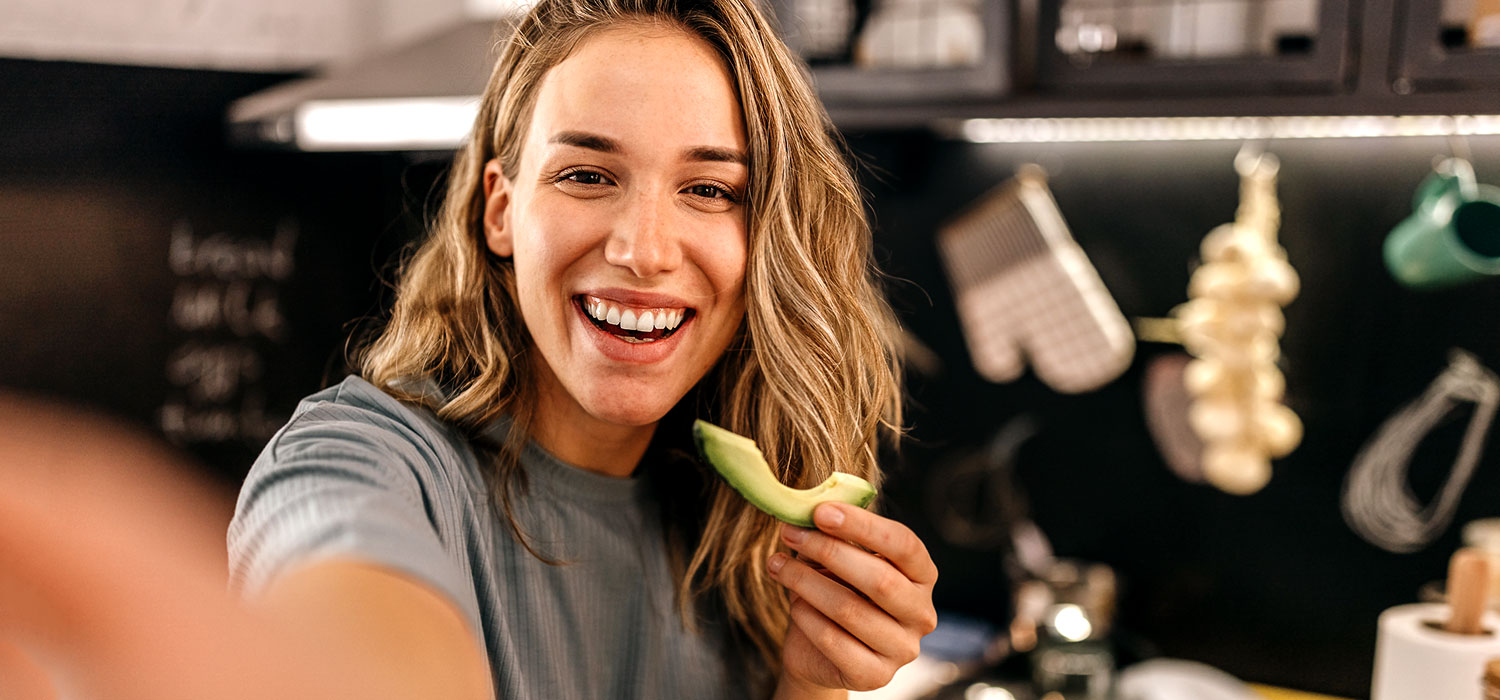7 Voedingsmiddelen die helpen om jonger te lijken Een jonge vrouw lacht terwijl ze een avocado eet