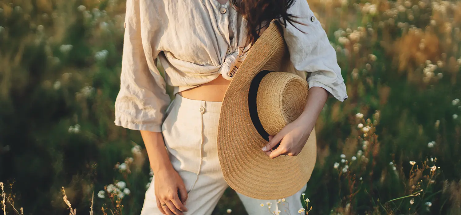 Onmisbare modetrends om je welzijn in 2025 te versterken Vrouw in een veld, met beige linnen kleding en een strooien hoed in haar hand