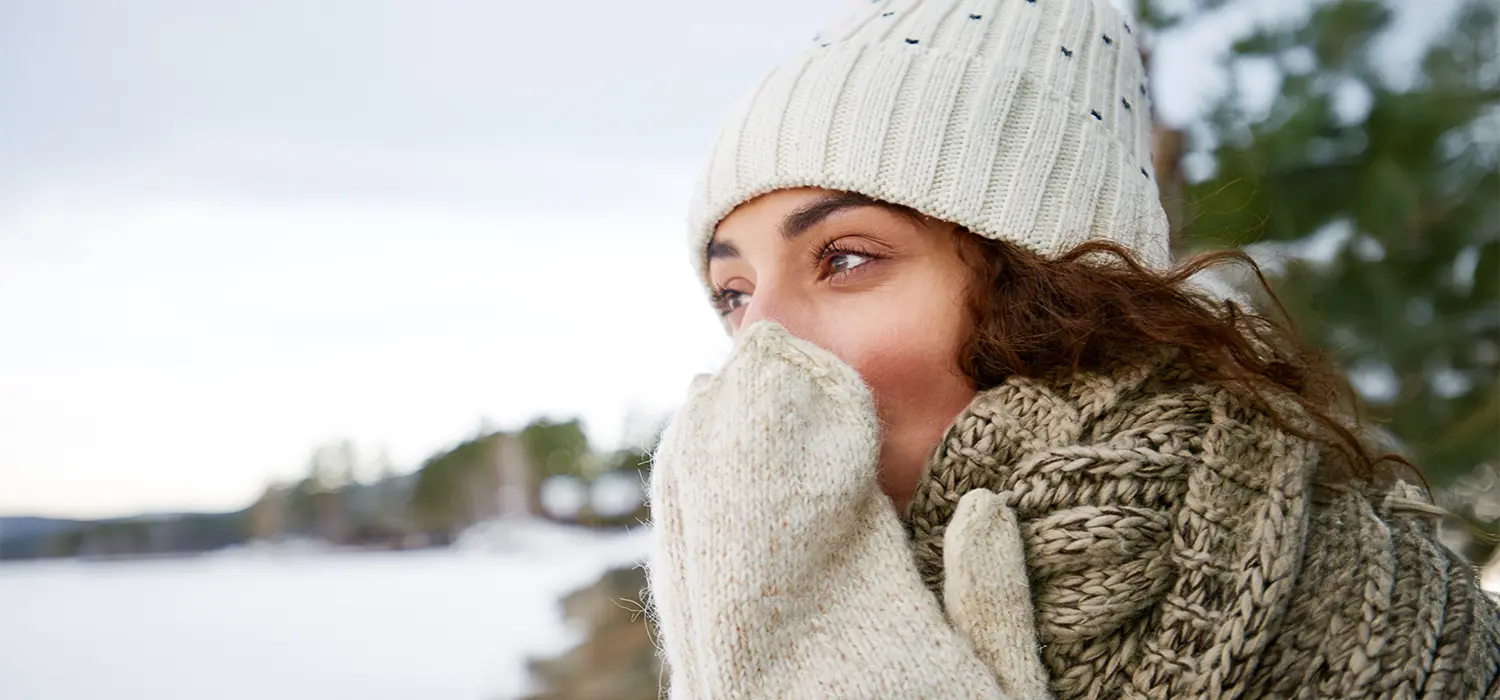 Waarom raakt de huidbarrière elke winter opnieuw beschadigd? Vrouw blaast in haar wanten buiten in een besneeuwd landschap